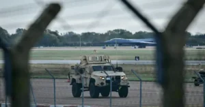 A military vehicle is seen on a runway with a large aircraft in the background, viewed through a fence.