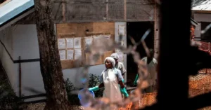 The photo shows a group of individuals in protective clothing and gloves working outside a building with a bulletin board, possibly in a rural or agricultural setting.