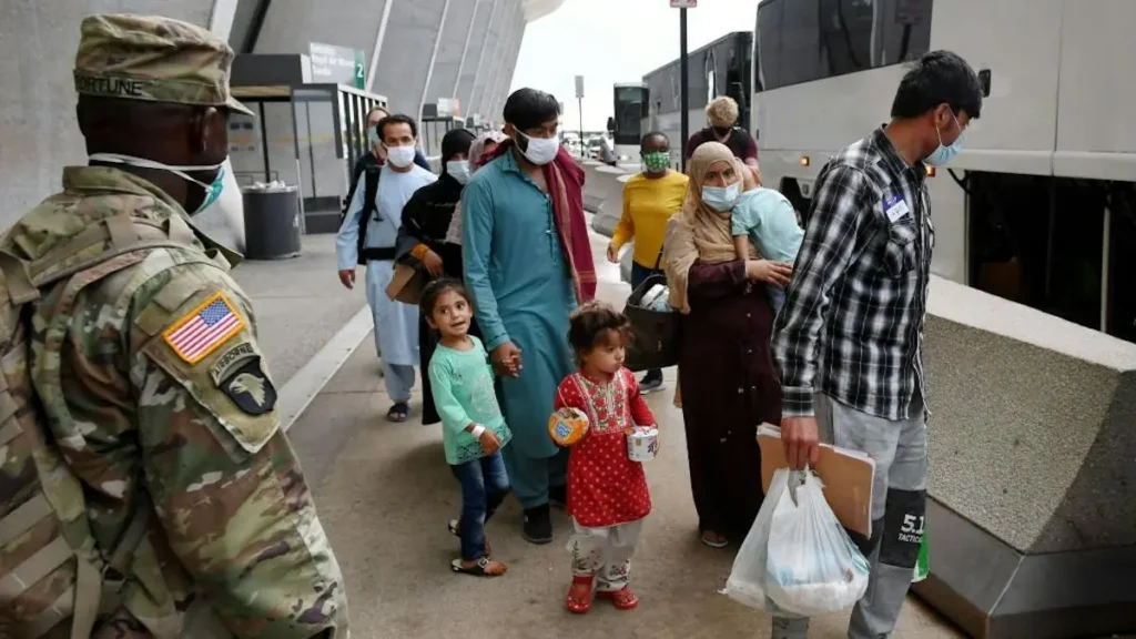 A group of people, including children, are seen walking near a bus stop, with some wearing masks and carrying bags, while a soldier in uniform stands nearby.