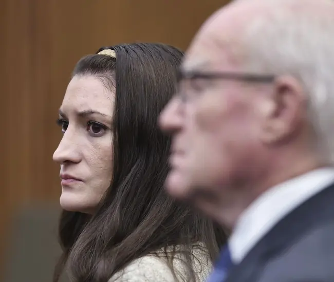The image shows a woman with long dark hair and a man in a suit, both appearing to be in a formal setting, possibly a courtroom or official event.