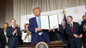 The image shows a person holding a signed document in front of a group of people, with a backdrop featuring the seal of the President of the United States and the text "America.gov."