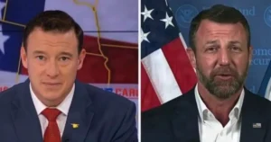 The image shows two individuals in formal attire, one wearing a suit with a red tie and the other in a suit with a white shirt, both set against a backdrop featuring an American flag and political imagery.
