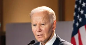 The image shows a man speaking at a podium with an American flag in the background.