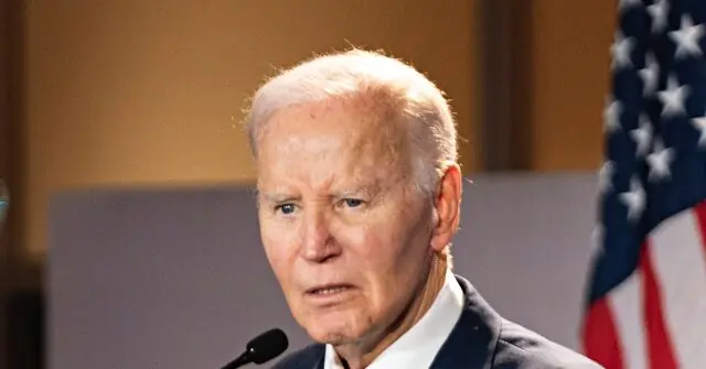 The image shows a man speaking at a podium with an American flag in the background.