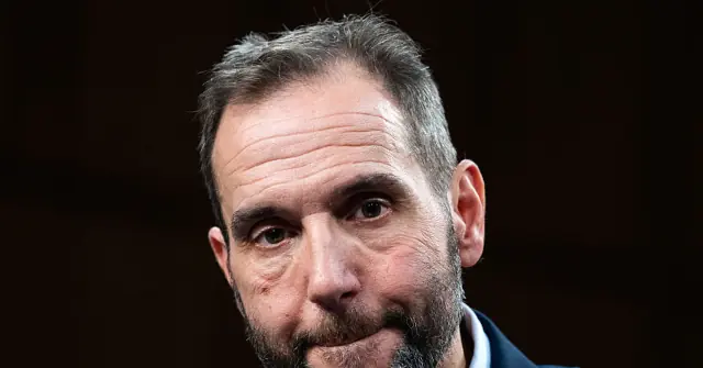 The image shows a close-up of a man with a beard and short hair, wearing a dark suit jacket.