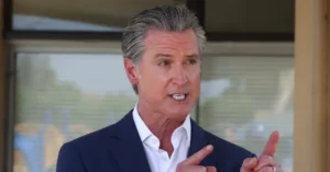 The image shows a man in a suit gesturing with his hands, likely speaking or explaining something.