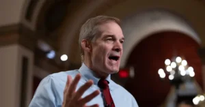 A man in a light blue shirt and red tie is speaking with his hand raised, likely during a formal or political event.