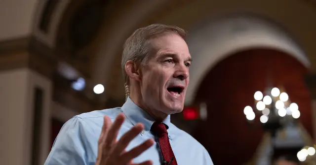 A man in a light blue shirt and red tie is speaking with his hand raised, likely during a formal or political event.