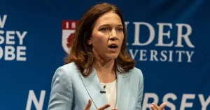 A woman is speaking at a podium in front of a backdrop featuring the "New Jersey" and "Rider University" logos.