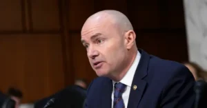 The image shows a man in a suit and tie speaking at a formal event, possibly a hearing or a meeting.