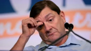 The image shows a man in a light blue shirt, appearing to be in a serious or contemplative mood, with his hand near his face.