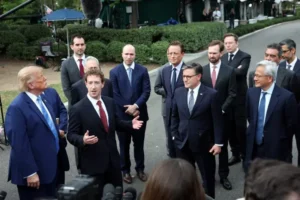 The photo shows a group of men in suits gathered outdoors, with one man speaking and others listening attentively.