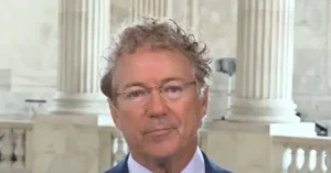 The image shows a man in a suit and tie, wearing glasses, standing in front of a backdrop of classical columns, likely in a formal or governmental setting.