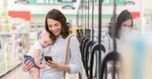 A woman smiles while holding a baby and looking at her phone in a store aisle.