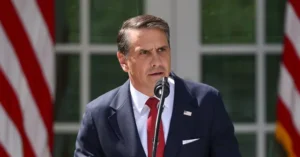 A man in a suit and tie stands at a podium with American flags in the background, delivering a speech.