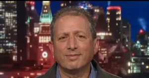 The image shows a person in a news broadcast setting, with a cityscape background featuring illuminated buildings.