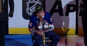 An elderly man in a hockey jersey plays a saxophone on stage during a ceremonial event.