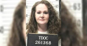 The image shows a mugshot of a person with long, curly hair, wearing a white shirt, and holding a sign with the text "TDOC 261368."