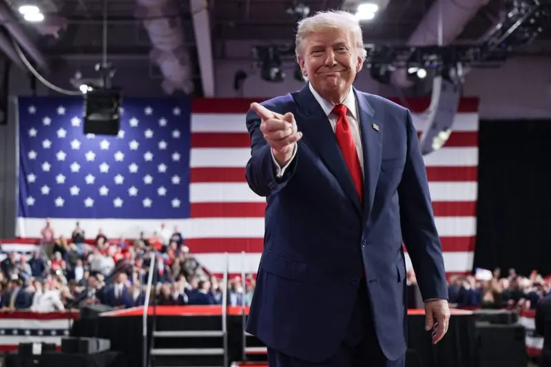 The image shows a person in a suit and red tie standing in front of an American flag, pointing towards the audience at a rally.