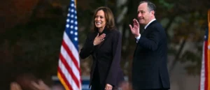 The image shows two individuals, one of whom is placing a hand over their heart, likely during a formal event or ceremony, with an American flag in the background.