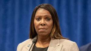 The image shows a person speaking at a press conference, with a microphone and a blue backdrop.