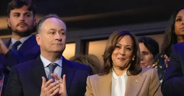 The photo shows a group of people, including a man in a suit and a woman in a beige blazer, clapping and smiling at an event.