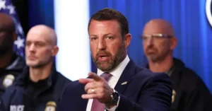 A man in a suit and tie is speaking at a press conference, pointing to the side, with two police officers in the background.