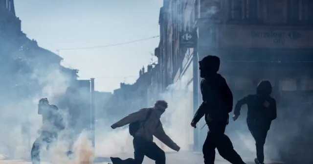 The image depicts a tense scene of protesters running through a cloud of tear gas in an urban setting.