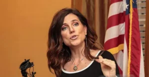 A woman is speaking in front of an American flag, gesturing with her hand.