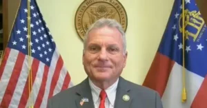 The image shows a man in a suit standing in front of the U.S. House of Representatives seal and two flags, one of which is the American flag.