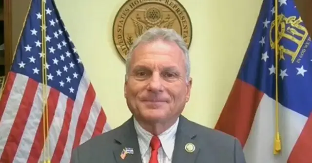 The image shows a man in a suit standing in front of the U.S. House of Representatives seal and two flags, one of which is the American flag.