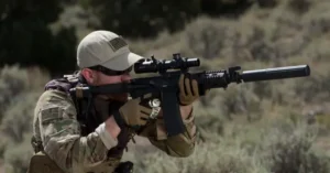 A soldier in camouflage attire aims a rifle equipped with a scope and suppressor in a desert environment.