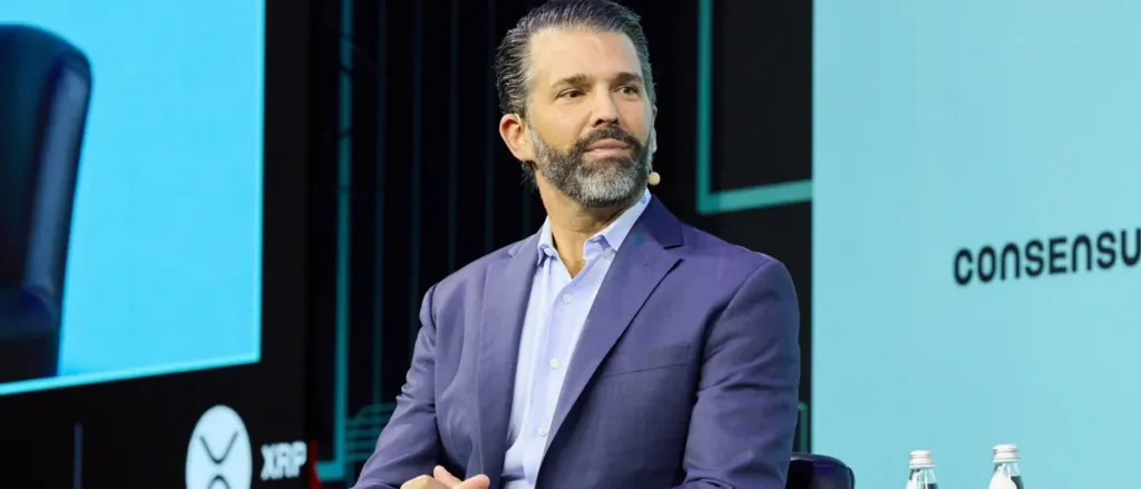 The image shows a man in a suit speaking at a conference, with a backdrop featuring the word "CONSENSUS" and logos for XRP.