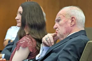 The photo shows two individuals seated in what appears to be a courtroom setting, with one person wearing a suit and the other in a red dress.