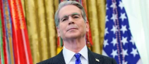 The image shows a man in formal attire standing in front of a backdrop featuring the American flag and other colorful banners.