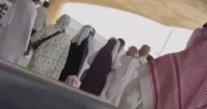 The image shows a group of people, some wearing traditional attire, gathered under a tent-like structure, possibly at a cultural or religious event.