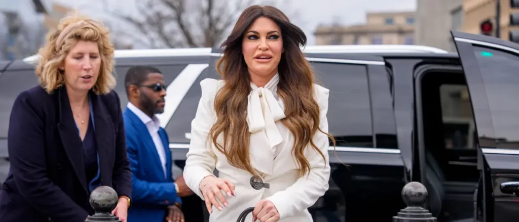 A woman in a white outfit with a bow at the neck is walking in front of a black limousine, accompanied by two other individuals.