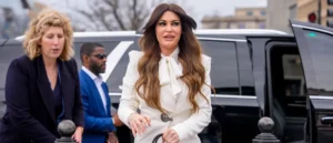 A woman in a white outfit with a bow at the neck is walking in front of a black limousine, accompanied by two other individuals.