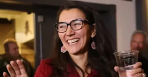 A woman in a red jacket and glasses is smiling and holding a drink, appearing to be in a social setting.