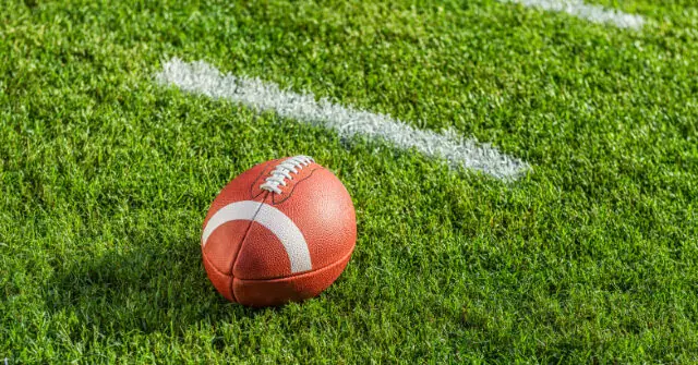 An American football rests on a grassy field marked with white yard lines.