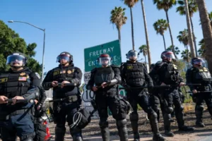 A line of police officers in riot gear stands in formation near a freeway entrance sign.