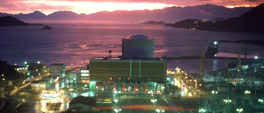 A nuclear power plant is illuminated at dusk, with a scenic view of the ocean and mountains in the background.