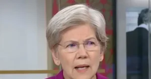 The image shows a person wearing glasses and a pink top, speaking in what appears to be a formal or professional setting.