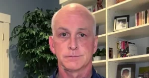 The image shows a man in a home office setting, with a bookshelf and a potted plant in the background.