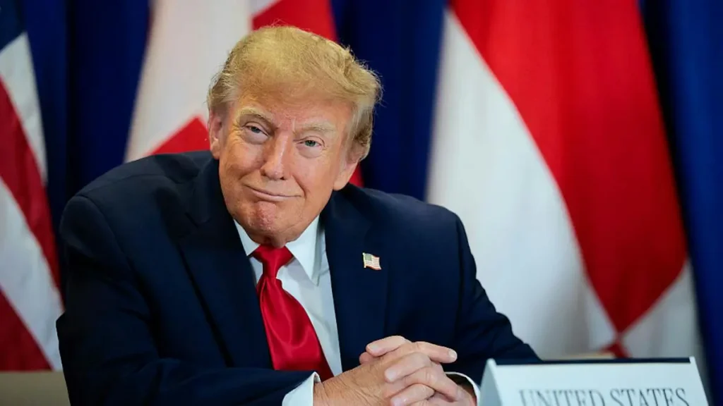 The image shows a person in a formal setting, wearing a suit and tie, with the United States flag visible in the background.