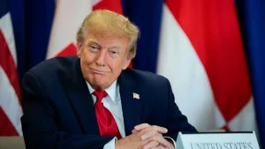 The image shows a person in a formal setting, wearing a suit and tie, with the United States flag visible in the background.
