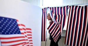 A man is seen inside a voting booth with an American flag in the background, symbolizing the voting process in the United States.