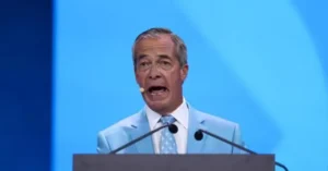 A man in a light blue suit and tie is speaking at a podium with microphones in front of a blue background.