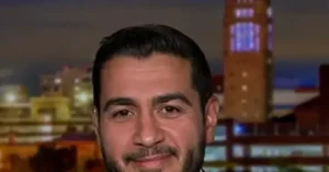 The image shows a man with a beard and short hair, smiling, with a cityscape and a tall tower in the background.