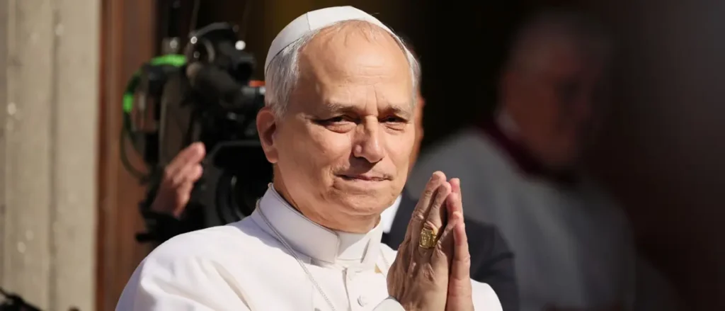The image shows a person in papal attire, with hands clasped in a gesture of prayer, standing in front of a camera setup.
